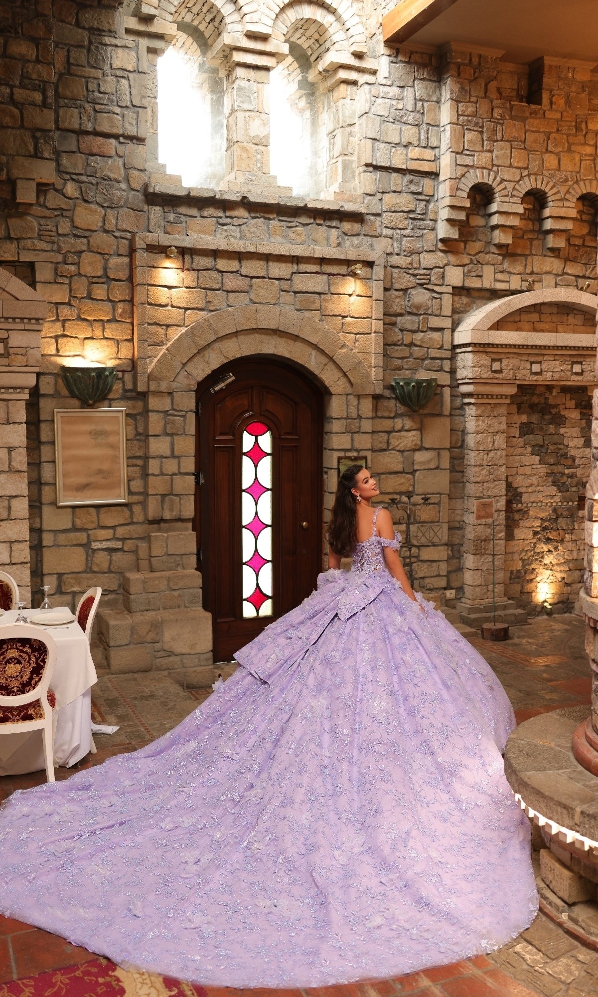 A woman in the Amarra 54243 off-shoulder quinceanera dress stands with her back to the camera in an ornate stone room, facing a wooden door with a stained glass window.